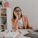 Uma mulher usando óculos e um blazer rosa está sentada em sua mesa, uma mão no rosto, parecendo entediada ou cansada. Em meio a cadernos espalhados e lápis de cor, ela contempla como aumentar a concentração no trabalho através do yoga enquanto prateleiras se avultam ao fundo. | YogaLounge
