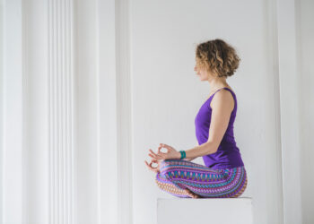 Uma pessoa com cabelo curto e cacheado está sentada de pernas cruzadas em uma superfície branca. Ela veste uma regata roxa e leggings coloridas, meditando com as mãos em um gesto de mudra contra uma parede branca, praticando yoga para controlar ataques de ansiedade. | YogaLounge