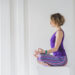 Uma pessoa com cabelo curto e cacheado está sentada de pernas cruzadas em uma superfície branca. Ela veste uma regata roxa e leggings coloridas, meditando com as mãos em um gesto de mudra contra uma parede branca, praticando yoga para controlar ataques de ansiedade. | YogaLounge