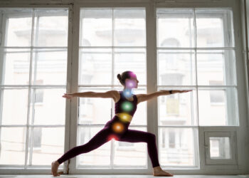 Em uma sala iluminada pelo sol, uma pessoa pratica ioga em uma pose de guerreiro, vestida com traje escuro. Uma ilustração vibrante de chakras, simbolizando energia fluindo, alinha-se da cabeça à pélvis. Grandes janelas iluminam o cenário sereno. | YogaLounge