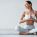 Uma mulher com um sutiã esportivo branco e leggings cinza-claro está sentada de pernas cruzadas em um tapete de yoga, irradiando calma. Com as mãos em posição de oração, olhos suavemente fechados, ela sorri gentilmente contra um fundo simples — incorporando a paz e o foco que vêm de deixar de fumar com yoga. | YogaLounge
