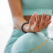 Uma pessoa senta-se de pernas cruzadas em uma pose de yoga, incorporando a essência de "Ter calma com o Yoga". Vestindo roupas esportivas azul-petróleo, sua mão repousa sobre o joelho em um gesto meditativo, adornada com uma pulseira de contas pretas. O fundo suavemente desfocado realça a cena serena. | YogaLounge