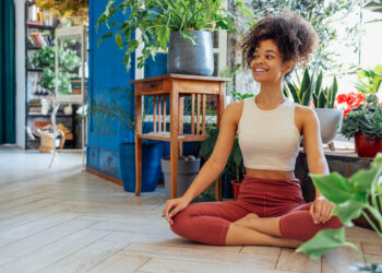 Uma pessoa com cabelo cacheado, top branco e calça vermelha está sentada de pernas cruzadas em um piso de madeira, sorrindo. Abraçando a felicidade através do yoga, eles estão cercados por várias plantas verdes em uma sala iluminada e aconchegante. | YogaLounge