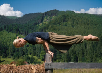 Uma pessoa com longos cabelos loiros presos em um rabo de cavalo se equilibra horizontalmente sobre as mãos em cima de uma cerca de madeira, fazendo uma pose de ioga ou ginástica, com colinas verdejantes e céu azul ao fundo. | YogaLounge