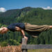 Uma pessoa com longos cabelos loiros presos em um rabo de cavalo se equilibra horizontalmente sobre as mãos em cima de uma cerca de madeira, fazendo uma pose de ioga ou ginástica, com colinas verdejantes e céu azul ao fundo. | YogaLounge