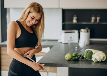 Em uma cozinha moderna, uma mulher em traje de ginástica sorri enquanto mede sua cintura com uma fita métrica. Vegetais frescos como maçã verde, brócolis e couve-flor sugerem sua jornada "Cardápio Para Emagrecer" espalhados pelo balcão ao lado dela. | YogaLounge