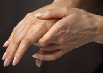 Close de duas mãos; uma segurando e apoiando delicadamente o dorso da outra, destacando a região do Canal Cárpico. A pele parece levemente enrugada, com veias e unhas naturais visíveis. O fundo é neutro e desfocado. | YogaLounge