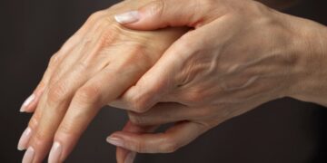 Close de duas mãos; uma segurando e apoiando delicadamente o dorso da outra, destacando a região do Canal Cárpico. A pele parece levemente enrugada, com veias e unhas naturais visíveis. O fundo é neutro e desfocado. | YogaLounge