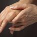Close de duas mãos; uma segurando e apoiando delicadamente o dorso da outra, destacando a região do Canal Cárpico. A pele parece levemente enrugada, com veias e unhas naturais visíveis. O fundo é neutro e desfocado. | YogaLounge