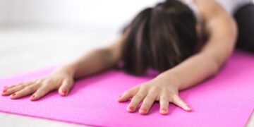 Uma pessoa com unhas pintadas se estica para frente sobre um tapete de ioga rosa brilhante, com os braços estendidos e a testa próxima ao tapete, praticando uma postura relaxante de ioga em ambientes fechados para ajudar a aliviar o estresse pós-traumático. | YogaLounge