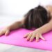 Uma pessoa com unhas pintadas se estica para frente sobre um tapete de ioga rosa brilhante, com os braços estendidos e a testa próxima ao tapete, praticando uma postura relaxante de ioga em ambientes fechados para ajudar a aliviar o estresse pós-traumático. | YogaLounge
