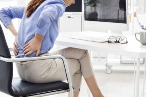 Uma mulher sentada em uma mesa sente dor na parte inferior das costas, o que sugere desconforto devido à má postura ao ficar sentada por muito tempo, com um computador, teclado e óculos na mesa à sua frente. | YogaLounge