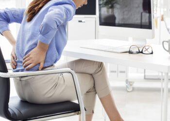 Uma mulher sentada em uma mesa sente dor na parte inferior das costas, o que sugere desconforto devido à má postura ao ficar sentada por muito tempo, com um computador, teclado e óculos na mesa à sua frente. | YogaLounge