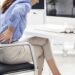 Uma mulher sentada em uma mesa sente dor na parte inferior das costas, o que sugere desconforto devido à má postura ao ficar sentada por muito tempo, com um computador, teclado e óculos na mesa à sua frente. | YogaLounge