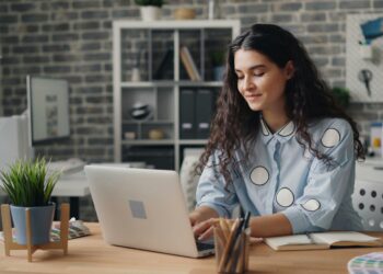 Uma mulher de cabelos longos e cacheados está sentada a uma mesa de madeira, digitando em seu laptop enquanto pesquisa um curso de ioga na Yoga Lounge Academy . Cadernos, canetas e uma planta estão sobre a mesa. Ela sorri levemente, com prateleiras e uma parede de tijolos ao fundo. | YogaLounge