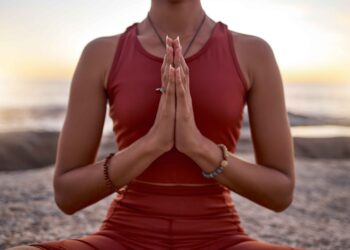 Uma pessoa vestindo uma roupa esportiva vermelha sentada de pernas cruzadas ao ar livre ao pôr do sol, segurando as mãos em posição de oração na frente do peito, praticando meditação ou ioga. | YogaLounge