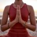 Uma pessoa vestindo uma roupa esportiva vermelha sentada de pernas cruzadas ao ar livre ao pôr do sol, segurando as mãos em posição de oração na frente do peito, praticando meditação ou ioga. | YogaLounge