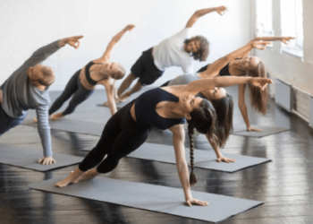 Um grupo de pessoas pratica uma postura de ioga de prancha lateral em tapetes em um estúdio iluminado durante um curso de ioga, todos estendendo um braço acima da cabeça e olhando na mesma direção. | YogaLounge