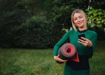 Uma mulher com roupa verde está ao ar livre na grama, segurando um tapete de ioga enrolado em um braço e olhando para o telefone com um leve sorriso — ela poderia ser uma Health Coach, cercada por árvores e arbustos. | YogaLounge