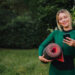 Uma mulher com roupa verde está ao ar livre na grama, segurando um tapete de ioga enrolado em um braço e olhando para o telefone com um leve sorriso — ela poderia ser uma Health Coach, cercada por árvores e arbustos. | YogaLounge