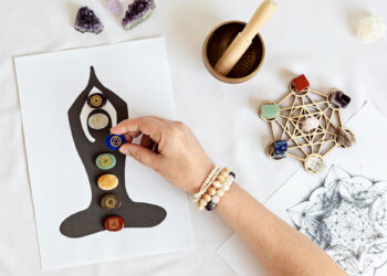 Uma mão coloca um cristal sobre um gráfico de chakras representando uma figura meditando. Vários cristais, contas e uma tigela de madeira com um martelo estão dispostos sobre uma superfície branca próxima. | YogaLounge