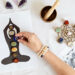 Uma mão coloca um cristal sobre um gráfico de chakras representando uma figura meditando. Vários cristais, contas e uma tigela de madeira com um martelo estão dispostos sobre uma superfície branca próxima. | YogaLounge
