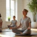 Cinco mulheres sentam-se com as pernas cruzadas em tapetes de ioga em uma sala iluminada pelo sol, praticando juntas a meditação MBSR. Elas usam roupas confortáveis e de tons neutros, e uma grande planta verde fica ao fundo, perto das janelas. | YogaLounge