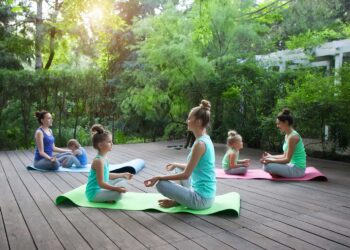 Três mulheres e três meninas praticam ioga em tapetes ao ar livre, ilustrando que não existe idade para fazer ioga. Elas sentam-se em pares em um deck de madeira, cercadas por uma vegetação exuberante, enquanto a luz do sol se filtra pelas árvores. | YogaLounge