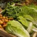 Uma caixa de madeira cheia de vegetais frescos, incluindo vagens, batatas pequenas e repolho chinês, fica ao ar livre, com terra e uma cesta de vime visíveis ao fundo. Health Coaching | YogaLounge