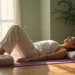 Uma mulher desfruta do Yoga Restaurativo em um tapete dentro de casa, deitada de costas com os joelhos dobrados, a cabeça sobre toalhas dobradas e uma almofada perto dos pés. A luz suave do sol entra pela janela e os vasos de plantas melhoram a atmosfera serena. | YogaLounge