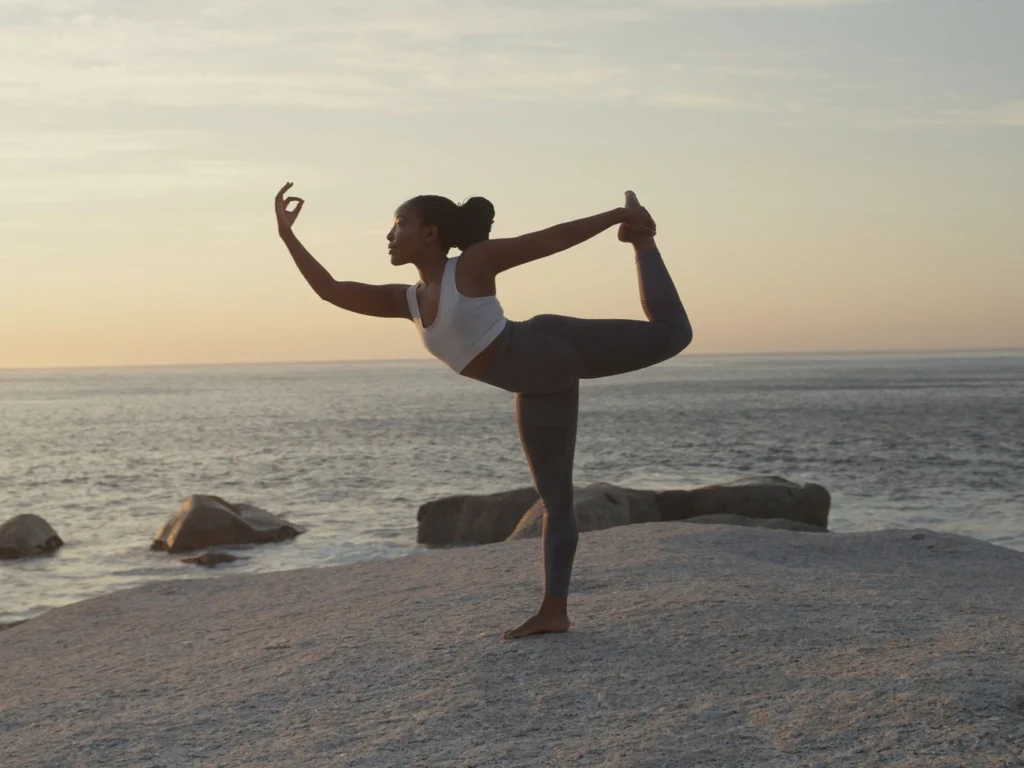 Uma mulher pratica uma postura de ioga à beira-mar ao pôr do sol, equilibrando-se em uma perna com a outra estendida atrás dela. Abraçando o Yoga no Verão, ela fica em uma superfície rochosa usando roupas esportivas. | YogaLounge