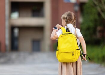 Uma jovem com duas tranças, vestindo camisa branca e saia bege, carrega uma mochila amarela brilhante e caminha para fora em direção a um prédio, segurando um chaveiro e usando um smartwatch. Ela está de costas para a câmera. | YogaLounge