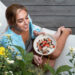 Uma mulher sorridente está sentada ao ar livre, perto de flores amarelas desabrochando, saboreando uma tigela de mingau de aveia com frutas vermelhas e nozes. Ela está descalça, com uma camisa azul clara, saboreando sua refeição - uma escolha perfeita para quem segue a Dieta Ayurvédica no Verão. | YogaLounge