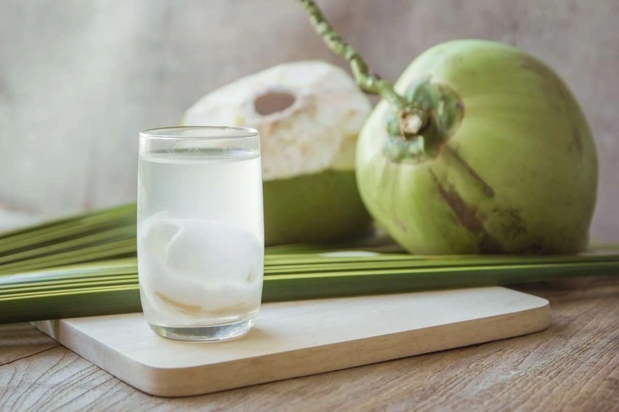 Um copo de água de coco com polpa de coco dentro fica em uma tábua de madeira, cercado por dois cocos verdes inteiros e folhas de palmeira - perfeito para hidratação no verão. | YogaLounge