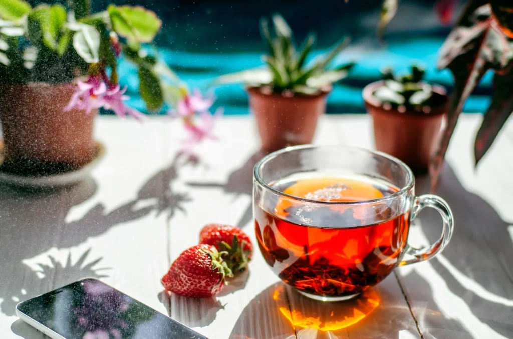 Uma xícara de chá de vidro está sobre uma mesa iluminada pelo sol com dois morangos, um smartphone e vasos de plantas ao fundo. A luz do sol e as sombras criam uma atmosfera aconchegante e fresca - perfeita para a hidratação no verão. | YogaLounge