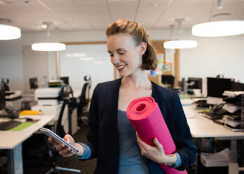 Mulher sorridente em traje de negócios segura um tapete de ioga rosa e olha para seu telefone em um escritório moderno com mesas e computadores ao fundo. | YogaLounge