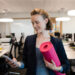 Mulher sorridente em traje de negócios segura um tapete de ioga rosa e olha para seu telefone em um escritório moderno com mesas e computadores ao fundo. | YogaLounge