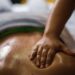 Um close-up de uma pessoa recebendo uma massagem ayurveda, mostrando uma mão pressionando suas costas oleadas. O foco é a mão e a pele relaxada e brilhante. | YogaLounge