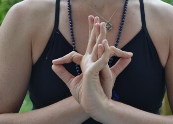Close-up de uma pessoa vestindo uma camiseta preta e um colar de contas, com as mãos unidas em um mudra de ioga na altura do peito, refletindo a conexão consciente praticada no ioga é ciência. O foco está nas mãos e na parte superior do tronco. | YogaLounge