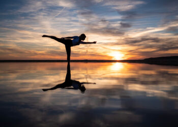 Uma pessoa pratica uma postura de ioga com uma perna só em uma superfície refletora ao pôr do sol, com nuvens dramáticas e cores vibrantes no céu espelhadas na água parada abaixo. | YogaLounge