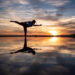 Uma pessoa pratica uma postura de ioga com uma perna só em uma superfície refletora ao pôr do sol, com nuvens dramáticas e cores vibrantes no céu espelhadas na água parada abaixo. | YogaLounge