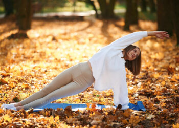 Uma mulher pratica uma postura de ioga com prancha lateral em um tapete azul ao ar livre, cercada por folhas de outono. Ela está usando um suéter branco e leggings bege, e sorri enquanto estica o braço para cima. | YogaLounge