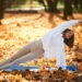 Uma mulher pratica uma postura de ioga com prancha lateral em um tapete azul ao ar livre, cercada por folhas de outono. Ela está usando um suéter branco e leggings bege, e sorri enquanto estica o braço para cima. | YogaLounge