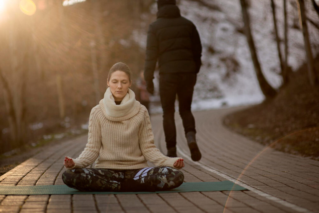 Uma mulher sentada de pernas cruzadas em um tapete de ioga meditando em um caminho pavimentado ao ar livre, usando um suéter creme. Uma pessoa com roupas escuras caminha ao fundo. A luz do sol brilha através das árvores. | YogaLounge