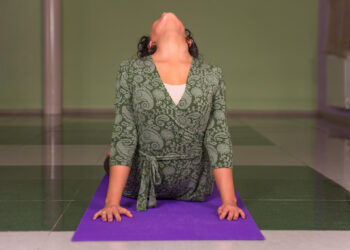 Uma pessoa com um top verde estampado pratica uma pose de ioga em um tapete roxo dentro de casa, arqueando as costas com a cabeça inclinada para cima e as mãos pressionando o tapete. | YogaLounge
