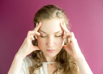 Uma mulher com cabelos longos e ondulados está de pé contra um fundo rosa, com os olhos fechados e os dedos pressionados nas têmporas, parecendo se concentrar ou aliviar o estresse - possivelmente refletindo sobre a mudança de hora e a ayurveda para o equilíbrio. | YogaLounge