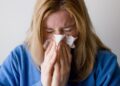 Uma pessoa de cabelos loiros vestindo um suéter azul está espirrando ou assoando o nariz em um lenço de papel, com os olhos bem fechados, possivelmente devido a um resfriado, gripe - que as técnicas de prevenção de gripe ayurveda podem ajudar a evitar. | YogaLounge