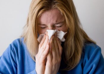 Uma pessoa de cabelos loiros vestindo um suéter azul está espirrando ou assoando o nariz em um lenço de papel, com os olhos bem fechados, possivelmente devido a um resfriado, gripe - que as técnicas de prevenção de gripe ayurveda podem ajudar a evitar. | YogaLounge