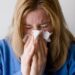 Uma pessoa de cabelos loiros vestindo um suéter azul está espirrando ou assoando o nariz em um lenço de papel, com os olhos bem fechados, possivelmente devido a um resfriado, gripe - que as técnicas de prevenção de gripe ayurveda podem ajudar a evitar. | YogaLounge