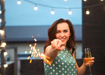 Uma mulher em um vestido verde de bolinhas sorri para a câmera enquanto segura um espumante e uma taça de champanhe, em pé ao ar livre sob luzes de corda à noite. | YogaLounge
